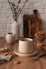 A ceramic cup with coffee, a milk jug, a glass bowl with cookies and a vase with branches on a...