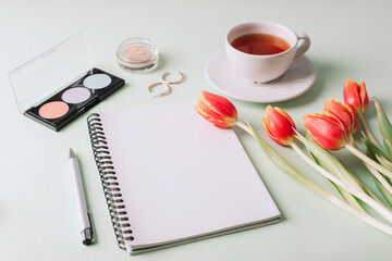 Blank notepad, cup of tea, makeup cosmetics and bouquet of red tulips on light green background. Top view, mockup. Women's day or Mother's day concept