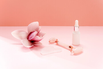 Obraz na płótnie Canvas Rose quartz crystal facial roller and gua sha scraper, face serum and magnolia flower on pink background. Facial massage kit for lifting therapy . Skin care anti-aging tools