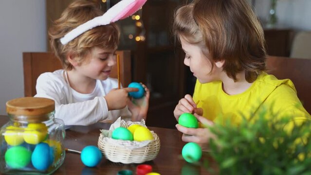 Easter Family traditions. Two caucasian happy siblings kids with bunny ears paint and decorate eggs with paints for holidays while sitting together at home table. Kids having fun together