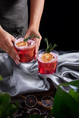 Red drink cocktail of rum and mezcal with lime and cherry juice garnished with dried fruit and green rosemary placed by two female hands