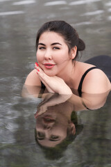 Extended sizes young woman in black swimming costume lying down geothermal mineral water in outdoors pool at balneotherapy spa, hot springs resort. Young adult model looking at camera. Body positive - obrazy, fototapety, plakaty