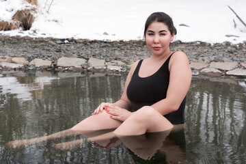 Outsize young adult woman in black one piece swimming costume sitting bathing in mineral water in outdoors pool at balneotherapy health spa. Femininity busty xxl woman looking at camera. Body positive - obrazy, fototapety, plakaty