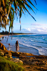 A beautiful girl in a dress stands on a rocky cliff looking at the famous Coolum beach and the Pacific Ocean and surfers catching big waves. Panorama from Sunshine Coast, Queensland, Australia