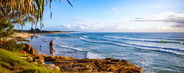 A beautiful girl in a dress stands on a rocky cliff looking at the famous Coolum beach and the Pacific Ocean and surfers catching big waves. Panorama from Sunshine Coast, Queensland, Australia - Powered by Adobe