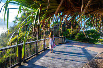 A girl in dress walks along the boardwalk and enjoys the view of the stunning Coolum Beach and surfers catching big waves, Pacific Ocean. Stunning panorama of Sunshine Coast, Queensland, Australia