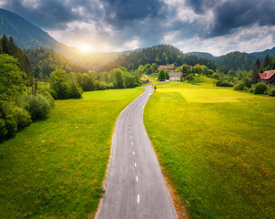 Aerial view of road in green meadows at sunset in summer. Top view from drone of rural road, mountains, forest. Beautiful landscape with roadway, sun rays, trees, hills, green grass, clouds. Slovenia