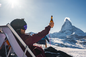 Young snowboarder cheering with a beer after skiing day in a bar or a cafe at the Zermatt ski...
