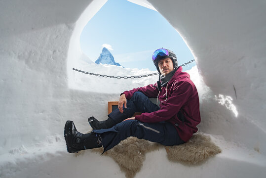 Portrait of smiling tourist snowboarder sitting inside an iglu dorf with a view on the famous snowcapped Matterhorn mountain. Relaxing in Swiss Alps, Zermatt ski resort.