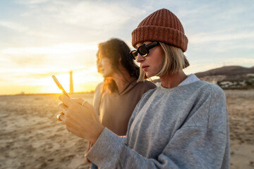Two girls portrait, one of them using mobile phone. Sunset time on the beach.