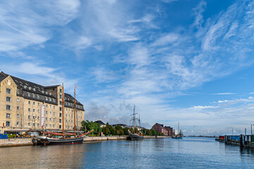 Fototapeta na wymiar Copenhagen, Denmark - September 13, 2010: Wide panorama with beige stone Admiral Hotel building along Larsens Place and behind Kvaesthusbroen quay with historic sailing yachts under blue cloudscape