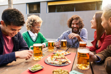 Multiracial friends having fun drinking beer together at terrace bar. Young people laughing at restaurant cafeteria. Happy guys and girls. High quality photo