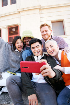 Vertical multiracial group selfie of smiling people. Young using cell phone outdoors. Cheerful boys and girls friends posing funny for photo. Happy vacations together. Social media and lifestyle.