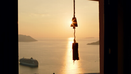 Windchimes hang on window, in which in background there is beautiful sunset over sea or ocean with...