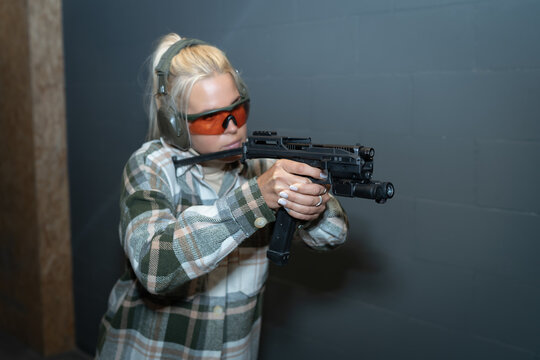 A beautiful girl in goggles and headphones takes aim from a tactical pistol in a shooting range.  Soft focus photo, main focus on the gun.