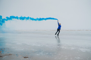 A caucasian woman is skating on a frozen lake holding colored blue smoke in her hands.
