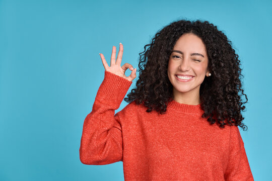 Young Happy Latin Woman Student Showing Ok Hand Okay Sign Winking Isolated On Blue Background. Smiling Female Model Advertising Sale Offer Presenting Promotion Recommending Discounts Concept.