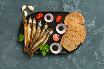 Board with delicious smoked capelin, bread pieces and vegetables on grey grunge background