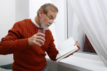 Upset senior man with glass of water reading book near window at home. Loneliness concept