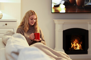 Fototapeta na wymiar Young woman with cup of hot drink resting near fireplace at home