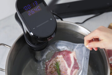Woman putting vacuum packed meat into pot with sous vide cooker in kitchen, closeup. Thermal...