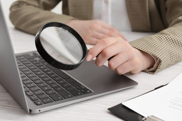 Woman holding magnifier near laptop at white wooden table, closeup. Online searching concept