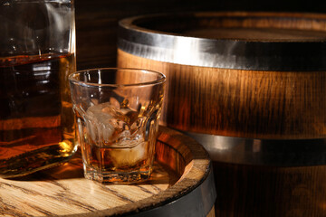 Glass of cold whiskey with oak barrels on wooden background