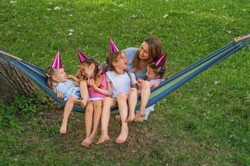 A mother rocks her daughters on hammock in the park and laughs merrily with them. Festive purple caps on the girls' heads. Children celebrate a friend's birthday.