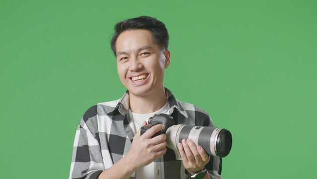 Close Up Of Asian Photographer Holding A Camera In His Hands And Smiling Being Happy While Standing On Green Screen Background In The Studio
