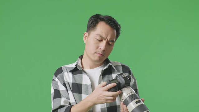 Close Up Of Asian Photographer Looking At Pictures In The Camera And Shaking His Head Being Unsatisfied With The Result On Green Screen Background In The Studio
