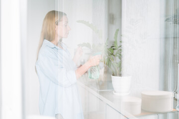 Young woman in blue shirt with spray with water in hands takes care of houseplant in kitchen at home through the window
