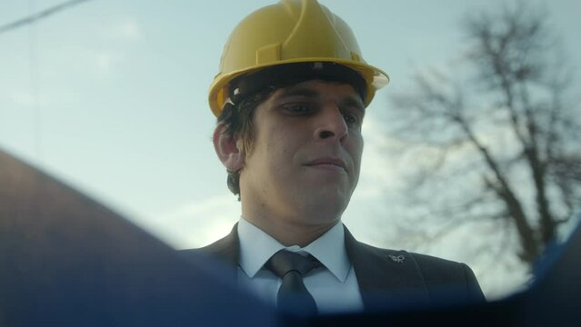 A Young, Beardless Construction Engineer or Contractor in a Helmet and Suit Examining Blueprints in a Blue Folder and Getting Frustrated over a Faulty Detail