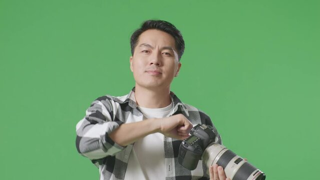 Close Up Of Asian Photographer Using A Camera Taking Pictures And Smiling Touching His Chest While Standing On Green Screen Background In The Studio
