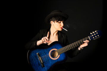 Obraz premium Young beautiful woman in black with a cigarette in her mouth holding an acoustic blue guitar