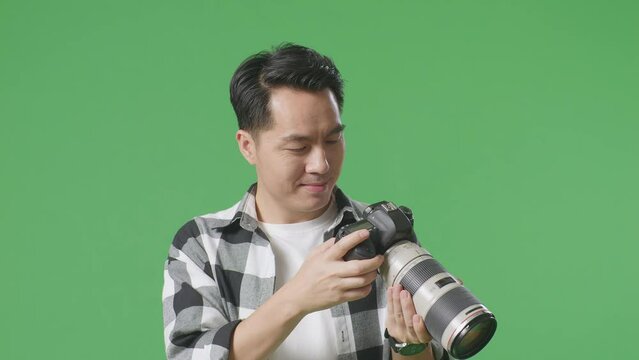 Close Up Of Asian Photographer Looking At The Pictures In The Camera And Smiling Being Satisfied With The Result While Standing On Green Screen Background In The Studio
