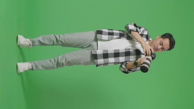 Full Body Of Asian Photographer Using A Camera Taking Pictures And Smiling Touching His Chest While Standing On Green Screen Background In The Studio
