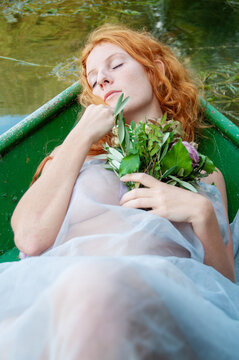beautiful longing sensual seductive young sexy redhead woman, Lady of Shalott with red hair, bride, lying with closed eyes and a bouquet of wilted flowers dreaming in a boat
