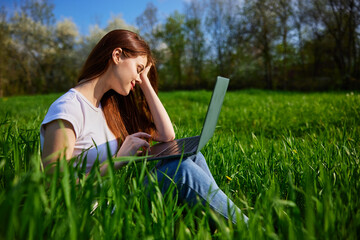 Young Caucasian woman using Laptop on nature, sitting in the grass in the park.