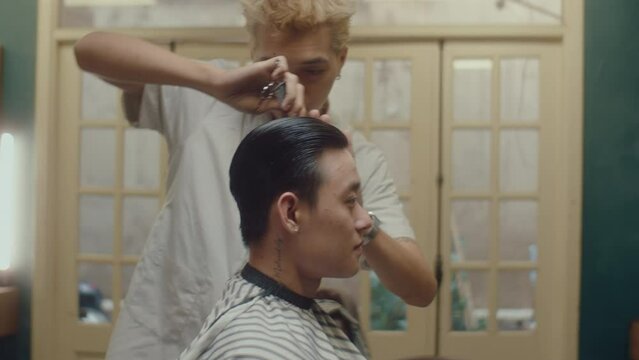 Young Asian man in cape getting new haircut in the barbershop, young barber combing his hair and chatting. Side view, medium shot