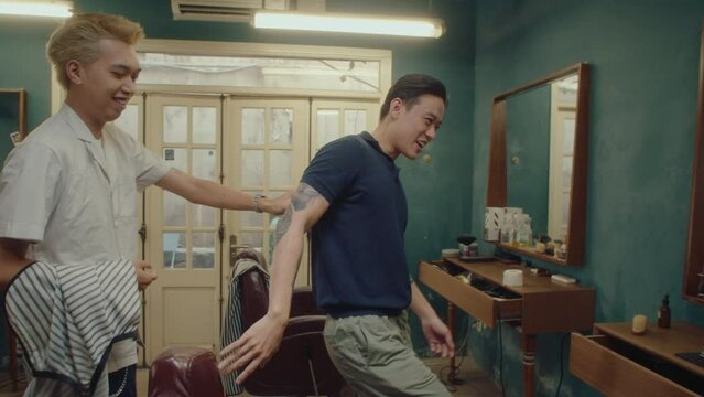 Young cheerful Asian barber greeting male customer at the barbershop. Man walking in the salon, sitting in chair and waiting for haircut