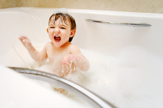 A happy child is playing with toys while sitting in a white homemade bathtub. Little toddler baby boy bathes in a bubble bath. Kid aged one year six months