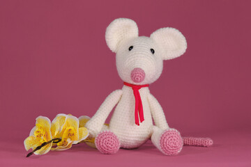 Cute little rat crocheted, handmade art. Amigurumi one white mouse with big ears and long nose sits on pink background next to yellow orchid flowers. A soft DIY toy made of natural cotton and wool.