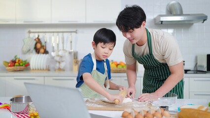 Obraz na płótnie Canvas Asian single father teaching little son making dough for bread, learning from videos on social media using labtop in the kitchen