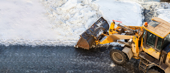 Orange tractor cleans up snow from the road and loads it into the truck. Cleaning of roads in the...