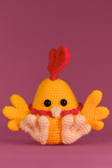 Orange cockerel with bright red comb on head and beak crocheted, handmade art. Front view. Amigurumi chicken doll on pink background. Soft DIY toy made of cotton and natural wool.