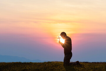 Silhouette Young male kneeling down with holding christian cross for worshipping God on the mountain sunset background.