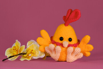 Amigurumi chicken doll on pink background with yellow orchid flowers. Soft DIY toy made of cotton and natural wool. Orange cockerel with bright red comb on head and beak crocheted, handmade art.