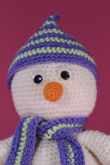 Soft DIY toy made of natural cotton and wool. Crocheted, handmade art. Amigurumi one small white snowman with orange nose and striped hat and scarf on pink background. Front view close portrait.