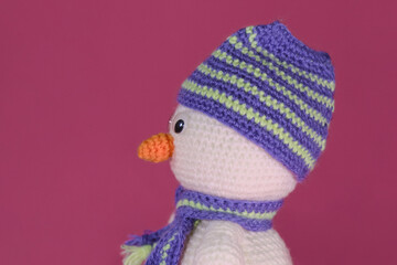 Soft DIY toy made of natural cotton and wool. Crocheted, handmade art. Amigurumi one small white snowman with orange nose and striped hat and scarf on pink background. Side view.