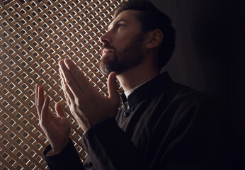 Catholic priest in cassock praying to God in confessional booth - obrazy, fototapety, plakaty